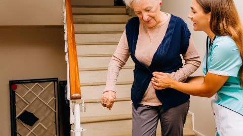 Une aide à domicile aide une personne âgée à descendre les marches d’un escalier d’immeuble.