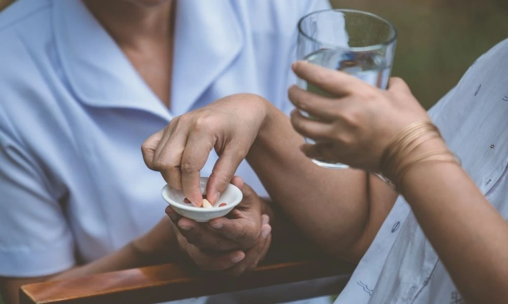 Une aide-soignante aide une personne à prendre ses médicaments en lui tendant un verre d’eau et une coupelle de comprimés.