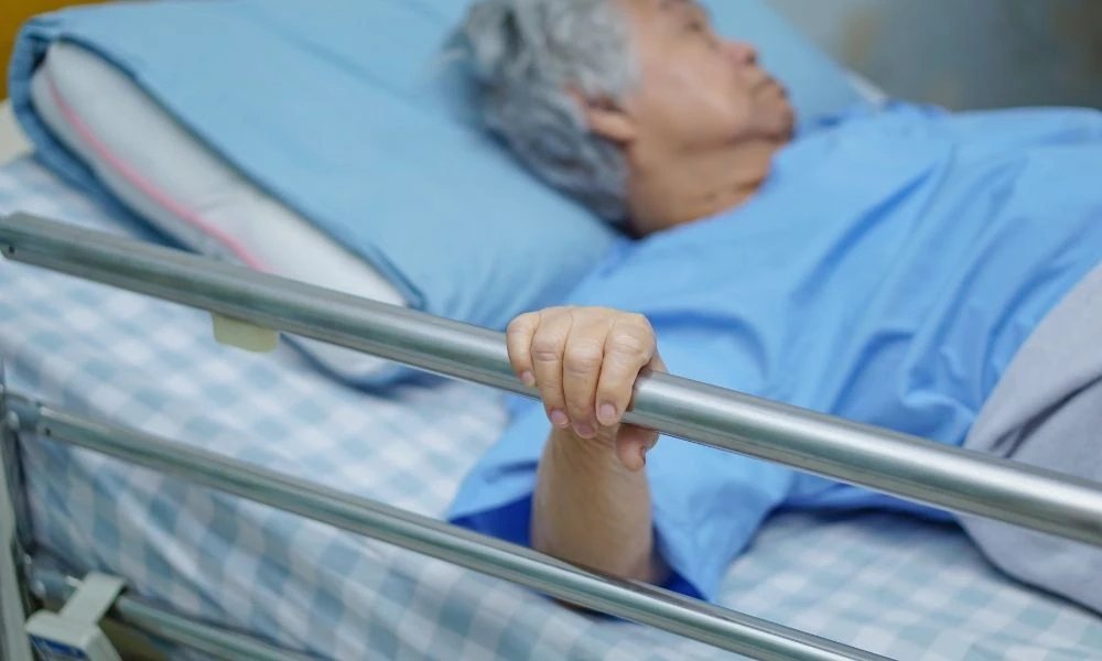 Une personne âgée est allongée dans un lit médicalisé tenant la barrière de sécurité du lit.