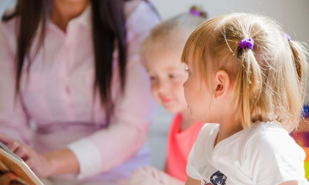 Deux jeunes enfants écoutent une adulte leur lire une histoire dans une ambiance douce et bienveillante.