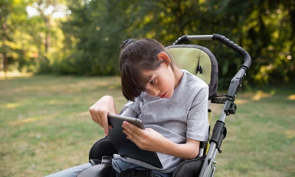 Une enfant en situation de handicap utilise une tablette tactile dans un parc, assis dans son fauteuil.