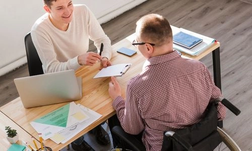 Un professionnel en fauteuil échange avec un collègue autour d’un dossier, dans une ambiance de travail collaborative et souriante.