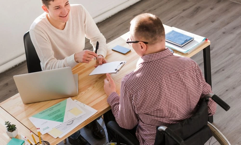 Un professionnel en fauteuil échange avec un collègue autour d’un dossier, dans une ambiance de travail collaborative et souriante.