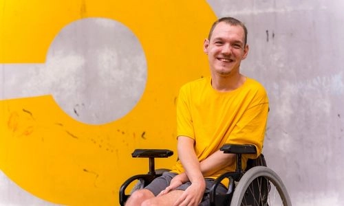 Un homme souriant, assis en fauteuil roulant, pose devant un grand mur jaune et gris au graphisme moderne.