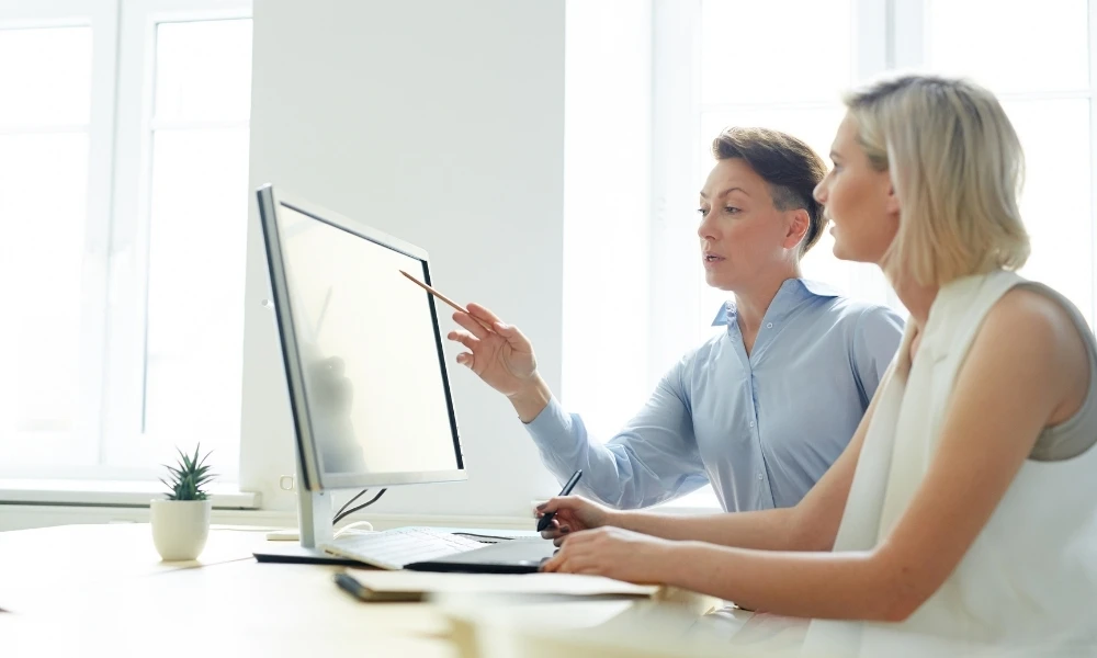 Deux femmes travaillent ensemble devant un ordinateur, l’une pointant l’écran avec un stylo.