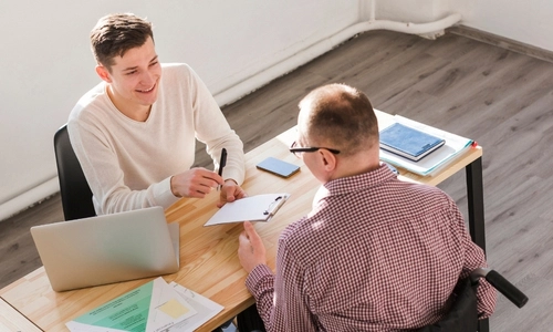 L'image montre deux personnes assises à un bureau en pleine conversation. L'une des personnes, à gauche, est un professionnel ou un conseiller, souriant et tenant un stylo, face à une autre personne à droite, qui est en fauteuil roulant.