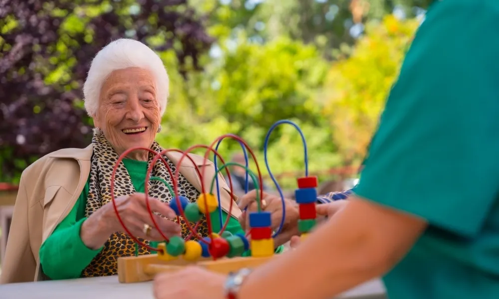L'image montre une personne âgée avec des cheveux blancs, assise à l'extérieur, souriant et interagissant avec un jeu de motricité en bois.