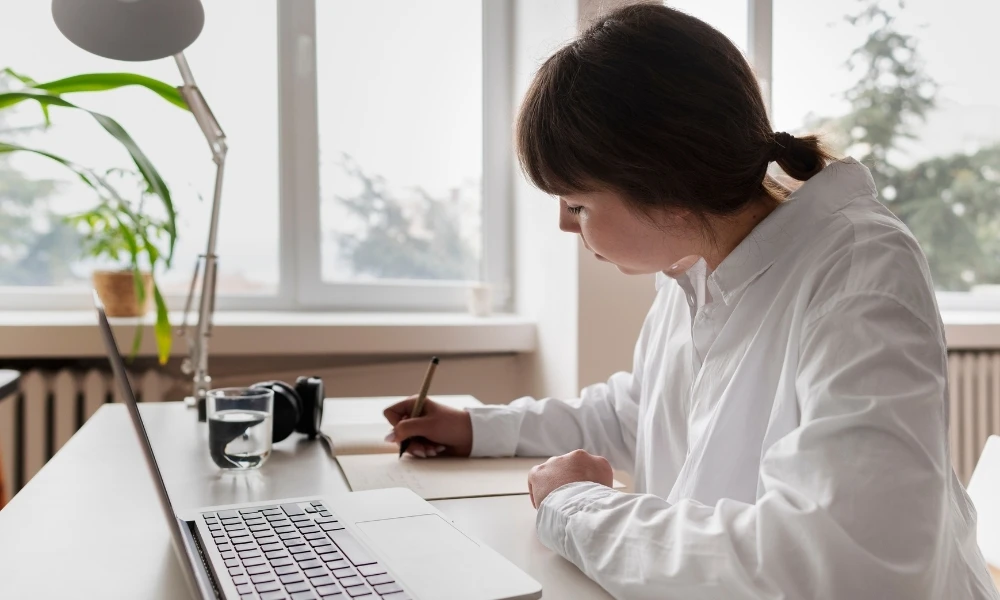 Cette image montre une jeune femme assise à un bureau, travaillant de manière concentrée. Elle écrit sur un carnet. Un ordinateur portable est ouvert sur le bureau devant elle.