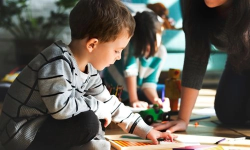 Un jeune enfant concentré joue au sol avec du matériel créatif, accompagné par un adulte dans un cadre calme et bienveillant.