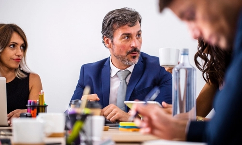 Scène d'une réunion professionnelle. Un homme en costume bleu, avec des cheveux poivre et sel et une barbe, est assis au centre de l'image. Il est en pleine conversation. À sa gauche (du côté droit de l'image), on distingue une femme avec de longs cheveux bruns, également concentrée sur la discussion.