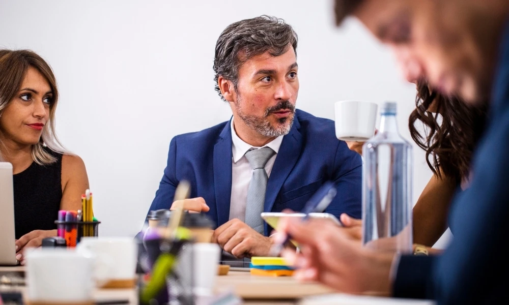 Scène d'une réunion professionnelle. Un homme en costume bleu, avec des cheveux poivre et sel et une barbe, est assis au centre de l'image. Il est en pleine conversation. À sa gauche (du côté droit de l'image), on distingue une femme avec de longs cheveux bruns, également concentrée sur la discussion.