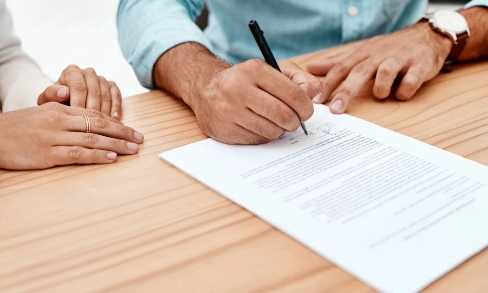 Deux personnes sont assises à une table en bois, l’une signant un document pendant que l’autre observe attentivement, suggérant une démarche contractuelle.