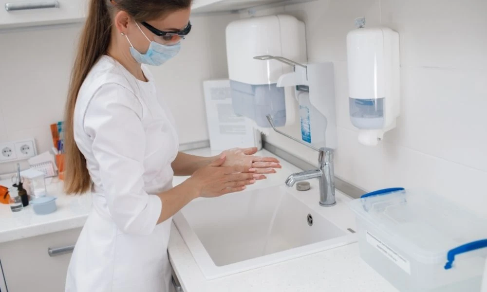 Une professionnelle de santé porte un masque et se lave soigneusement les mains au lavabo dans un environnement de soins.