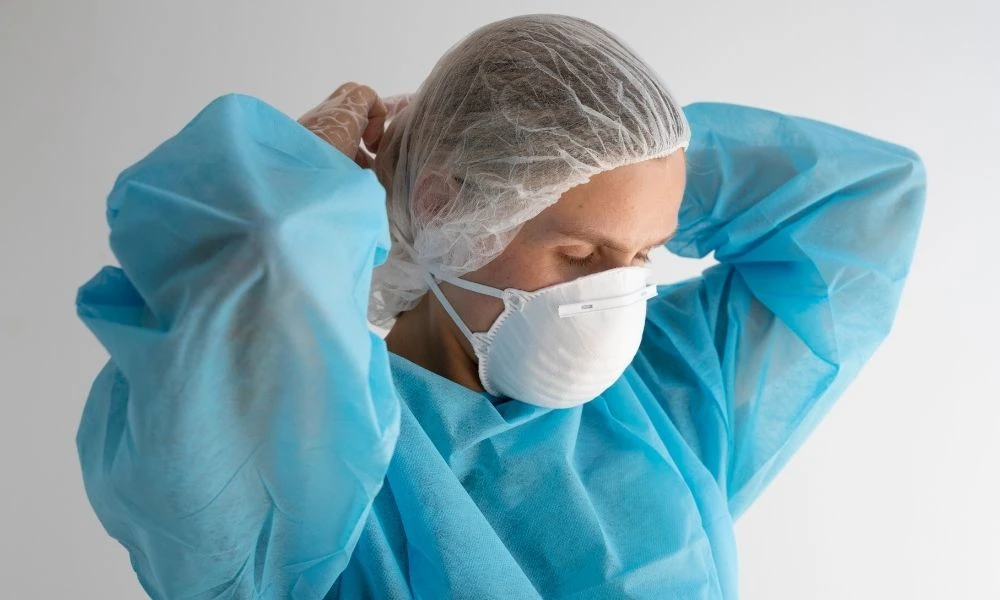 Un professionnel de santé ajuste son masque et sa charlotte, vêtu d’une surblouse bleue, en préparation pour des soins.