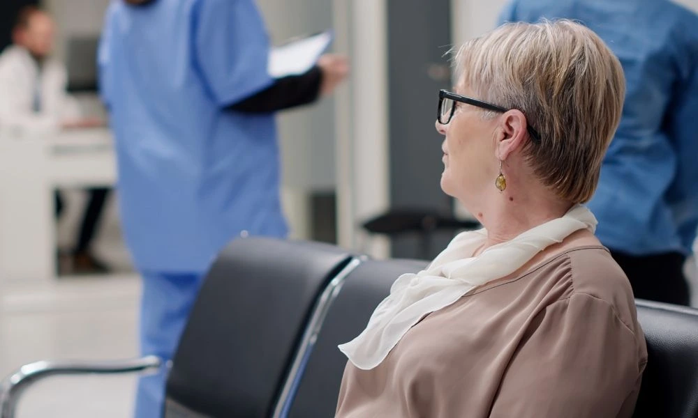 Une femme âgée, assise dans une salle d’attente médicale, observe le personnel soignant en arrière-plan.