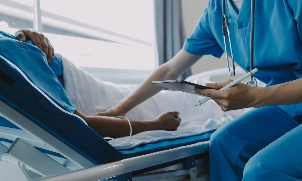 Un soignant en tenue médicale prend des notes au chevet d’un patient allongé dans un lit médicalisé.