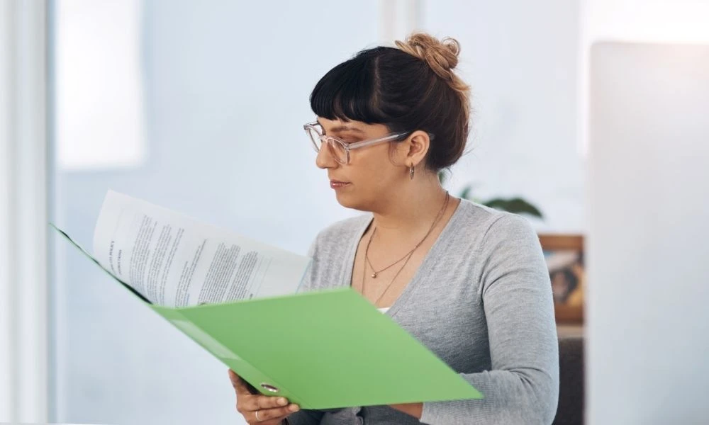 Une femme portant des lunettes lit attentivement un dossier dans un environnement calme et lumineux, évoquant un contexte professionnel.