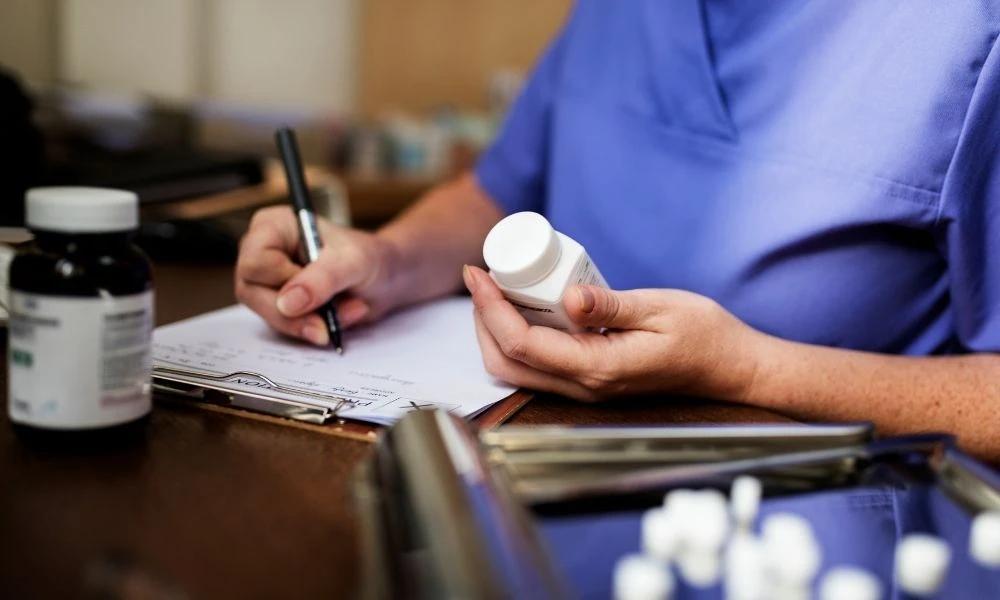 Un professionnel de santé en blouse bleue note des informations sur un dossier tout en tenant un flacon de médicaments.