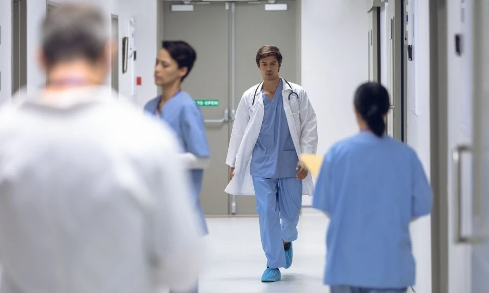 Un médecin en blouse blanche marche dans un couloir d’hôpital, entouré de soignants en tenue bleue, dans une ambiance clinique et professionnelle.