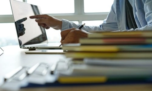 Une personne pointe l’écran d’un ordinateur portable tout en prenant des notes, avec des piles de documents floues au premier plan.