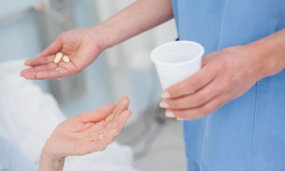 Un professionnel de santé en tenue bleue remet des comprimés et un verre d’eau à un patient prêt à les prendre.