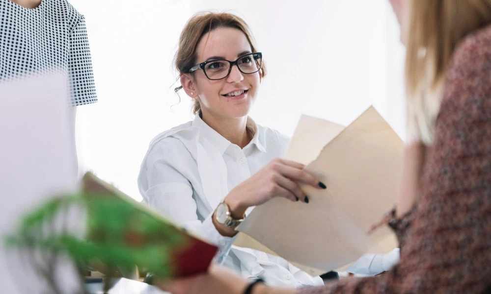 Une femme avec des lunettes remet des documents à une autre personne lors d’un entretien ou d’une réunion dans un bureau.