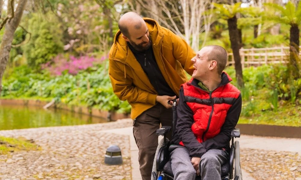 Deux hommes sont dans un parc ou un jardin.L'homme en fauteuil roulant sourit largement en regardant vers le haut vers l'autre homme qui est debout à l’arrière du fauteuil roulant.