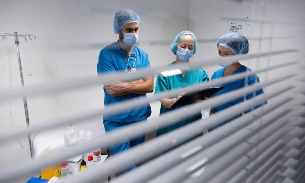 Trois professionnels de santé, vus à travers des stores, consultent un dossier médical dans une salle de soins.