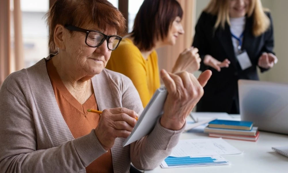 Une personne âgée écrit sur un carnet lors d’une réunion, tandis que deux professionnelles échangent en arrière-plan.