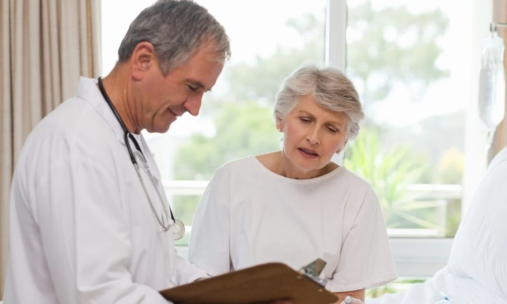 Un médecin montre un document sur un clipboard à une femme âgée dans une pièce lumineuse lors d’une consultation.