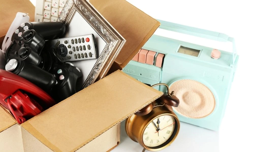 Une boîte en carton remplie d’objets variés (manette de jeu, réveil, téléphone, cadre) posée à côté d’une radio vintage.