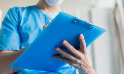 Un professionnel de santé en tenue médicale et masque tient un clipboard bleu et prend des notes.