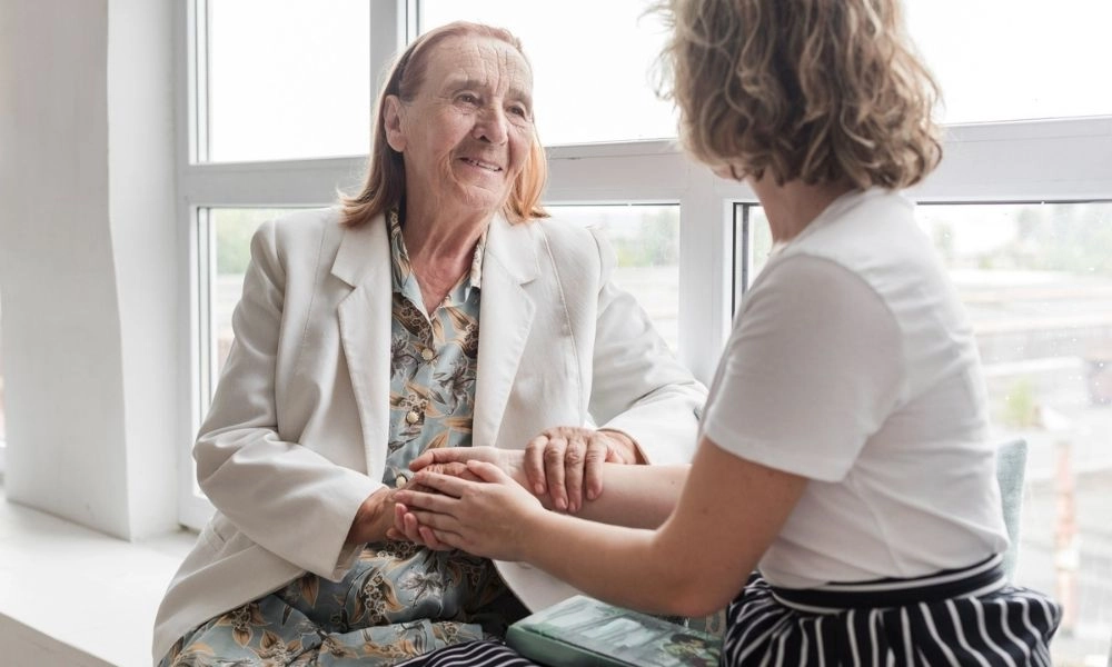 Une dame regarde avec bienveillance une personne âgée assise près d’une fenêtre, en lui tenant les mains.