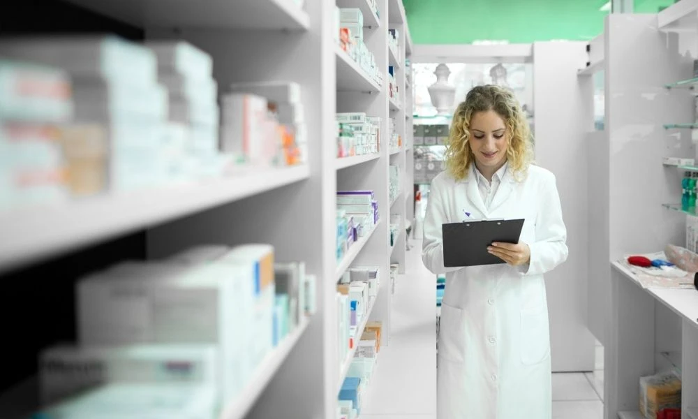 Une professionnelle de santé en blouse blanche vérifie des médicaments sur une tablette au milieu des rayonnages d’une pharmacie.