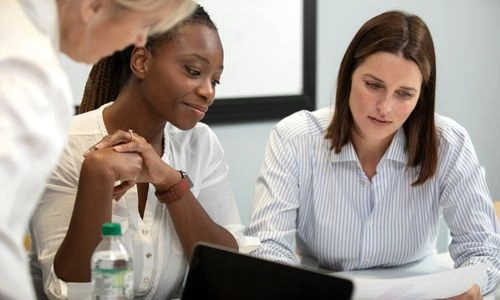 L'image montre trois femmes en train de discuter ensemble autour d'une table dans un cadre de travail ou de réunion.