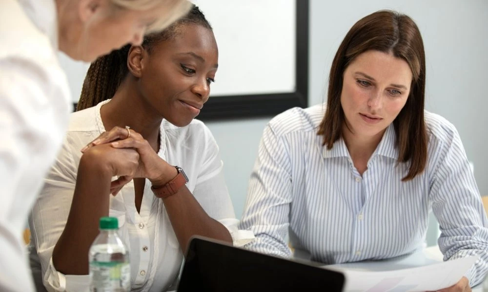 L'image montre trois femmes en train de discuter ensemble autour d'une table dans un cadre de travail ou de réunion.