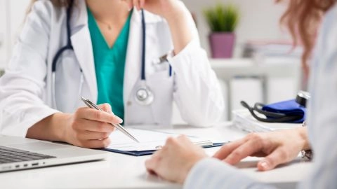 Une professionnelle de santé en blouse, stéthoscope autour du cou, prend des notes lors d’un échange avec une patiente assise en face d’elle.