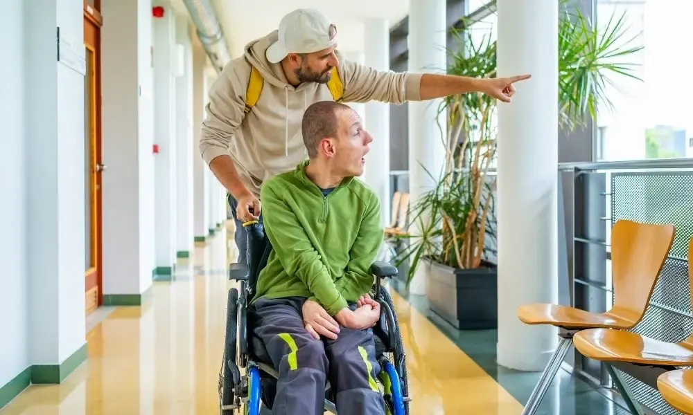 Un accompagnant pousse une personne en fauteuil roulant dans un couloir lumineux, tandis qu’ils regardent ensemble vers l’extérieur avec curiosité.