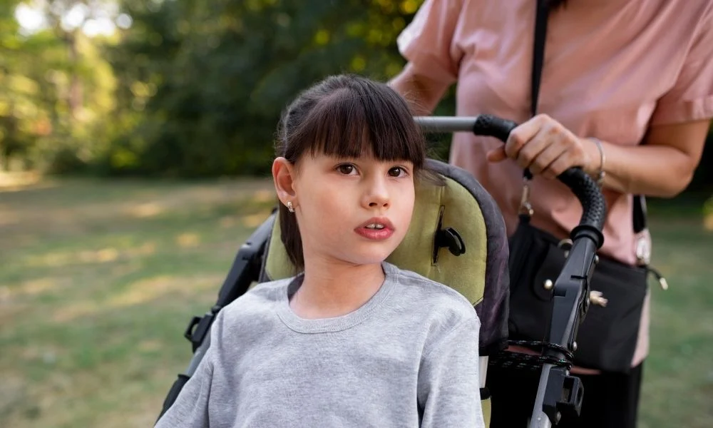 L'image montre un gros plan d'une jeune fille assise dans un fauteuil roulant, poussée par une femme en arrière-plan.