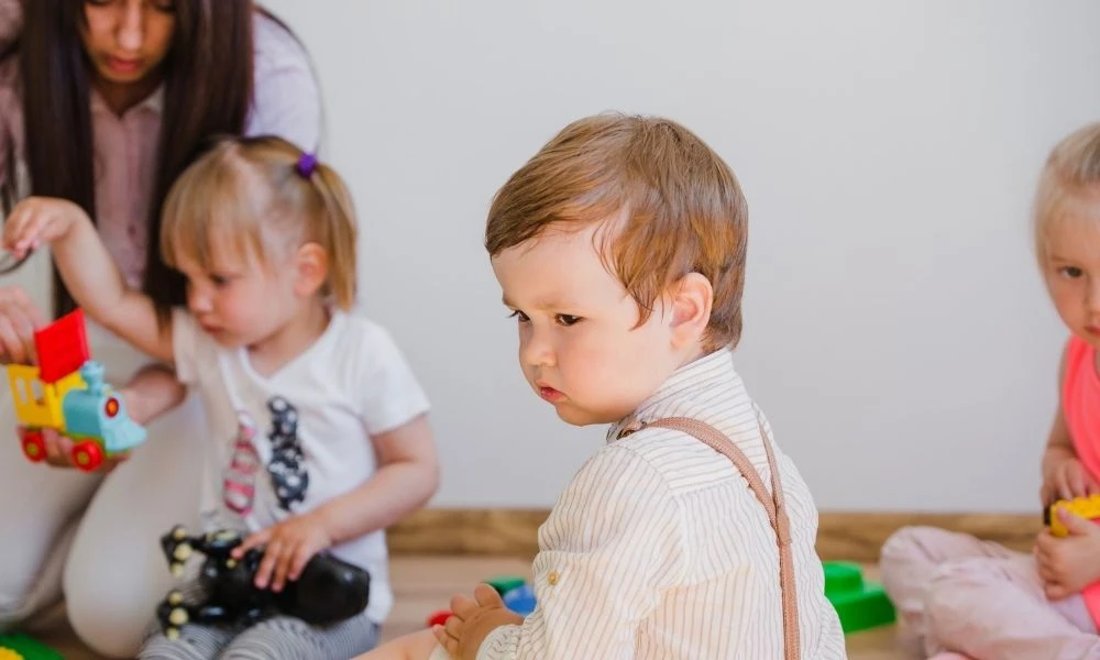Une professionnelle accompagne de jeunes enfants dans une activité de jeu en crèche, tandis qu’un tout-petit assis au premier plan semble observateur.
