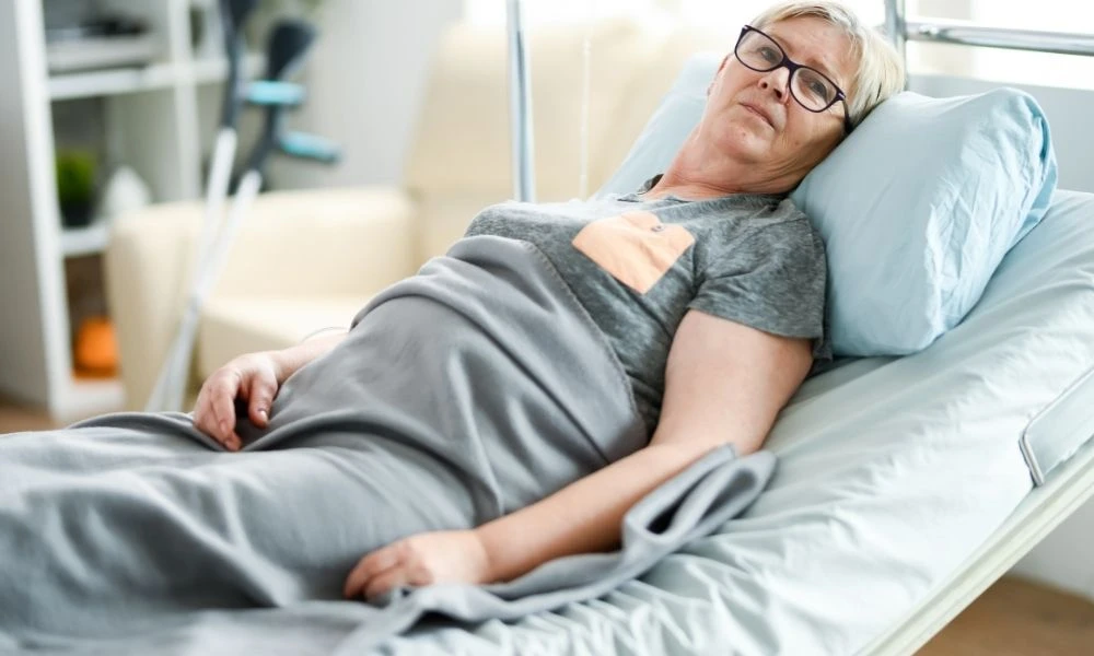 Une femme âgée est allongée dans un lit médicalisé chez elle, elle est couverte d'une couverture grise et semble se reposer.