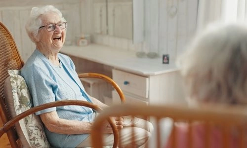 Femme âgée souriante assise dans un fauteuil à bascule, en conversation avec une autre personne dans un environnement intérieur lumineux.