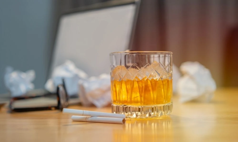 Un verre de whisky et deux cigarettes posés sur un bureau encombré, évoquant stress et addictions en milieu professionnel.