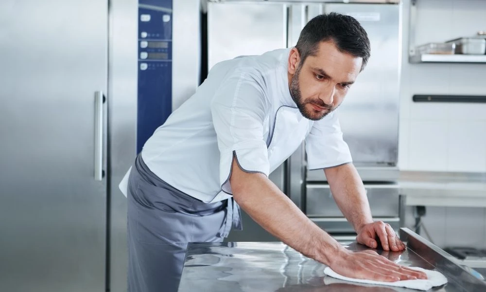 Un professionnel de restauration collective nettoie minutieusement un plan de travail en inox dans une cuisine professionnelle.