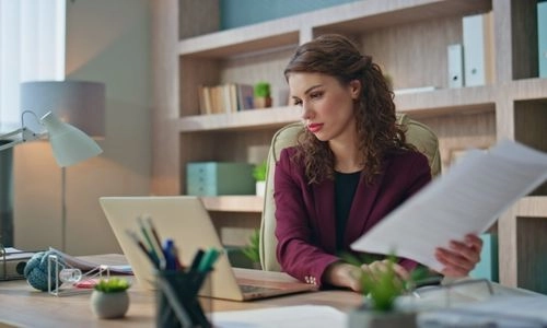 Une femme consulte des documents papier tout en travaillant sur son ordinateur portable dans un bureau chaleureux.