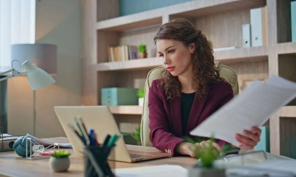 Une femme consulte des documents papier tout en travaillant sur son ordinateur portable dans un bureau chaleureux.