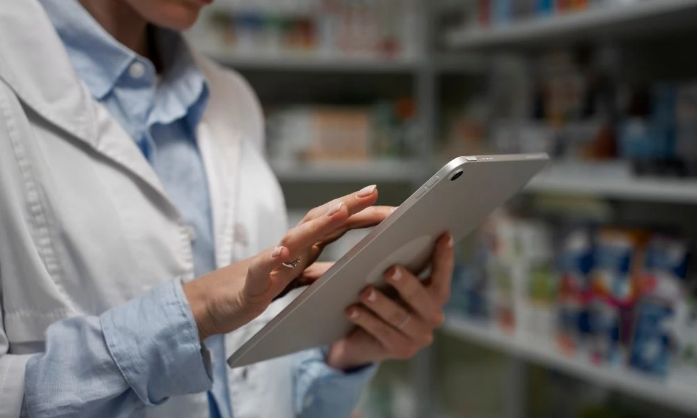 Pharmacienne consultant une tablette numérique dans une pharmacie, devant des rayonnages de médicaments.