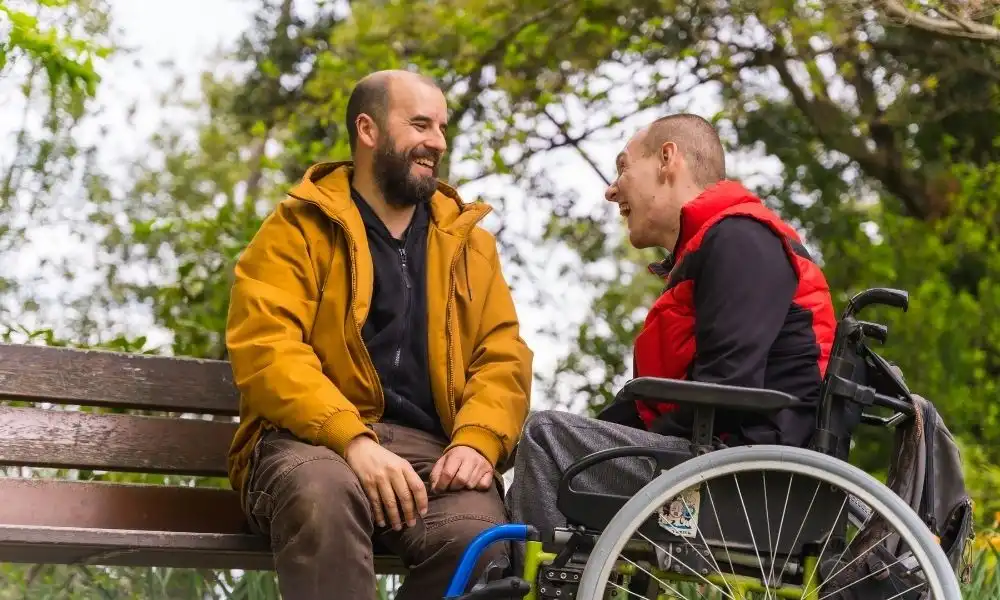 Deux hommes souriants échangent sur un banc dans un parc, l’un assis et l’autre en fauteuil roulant.