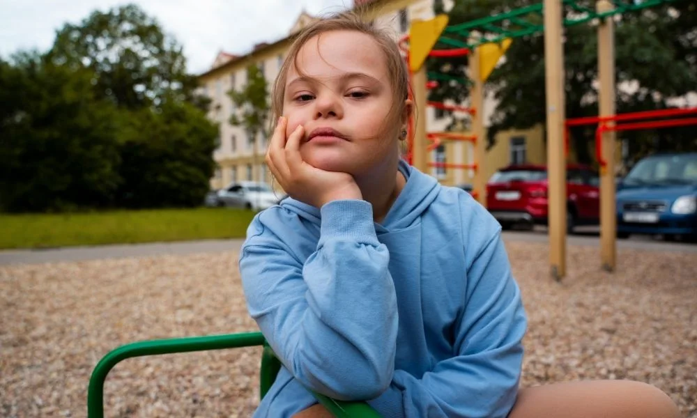 Petite fille atteinte de trisomie 21 assise sur un équipement de jeu dans un parc. Elle semble songeuse, appuyant sa tête sur sa main gauche avec un regard sérieux ou pensif.