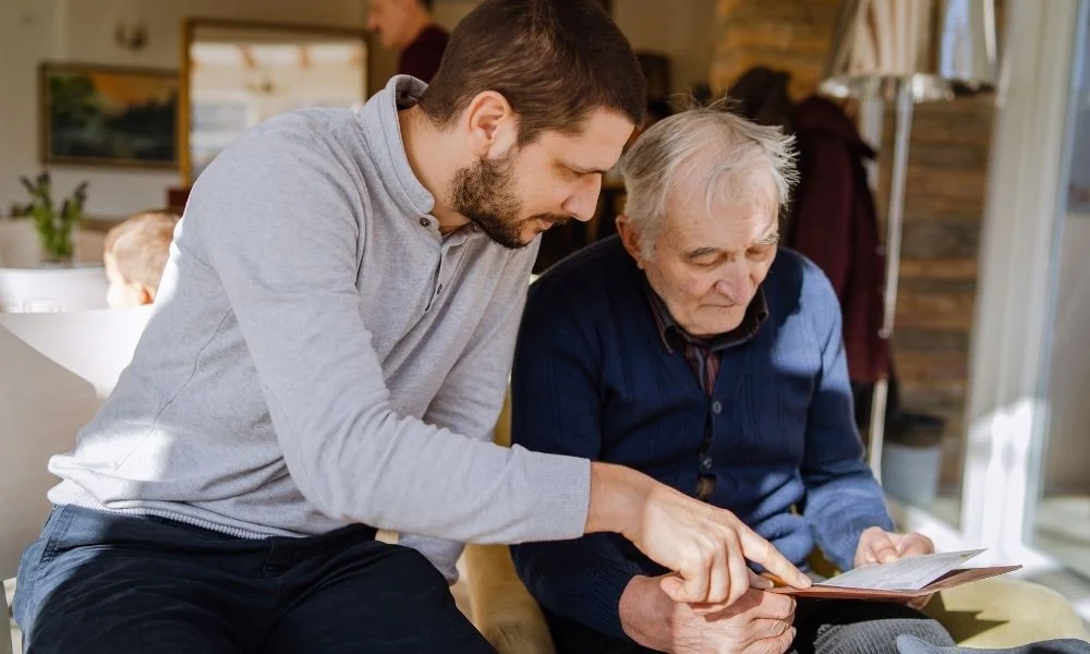 Un homme plus jeune accompagne une personne âgée en lui expliquant un document posé sur la table.